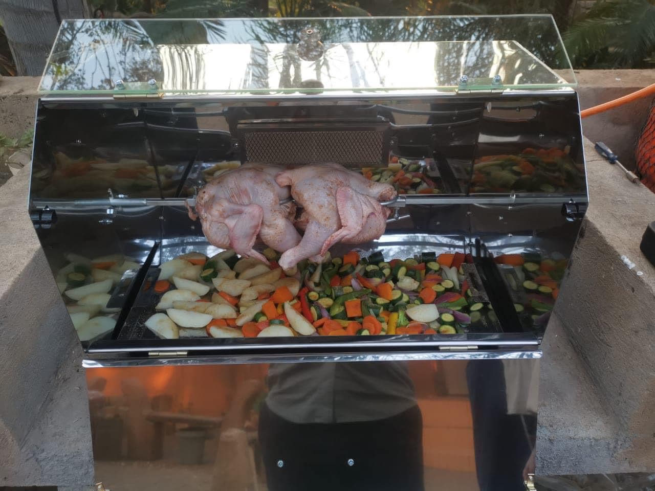 Two chickens on the spit rod above a full tray of vegetables