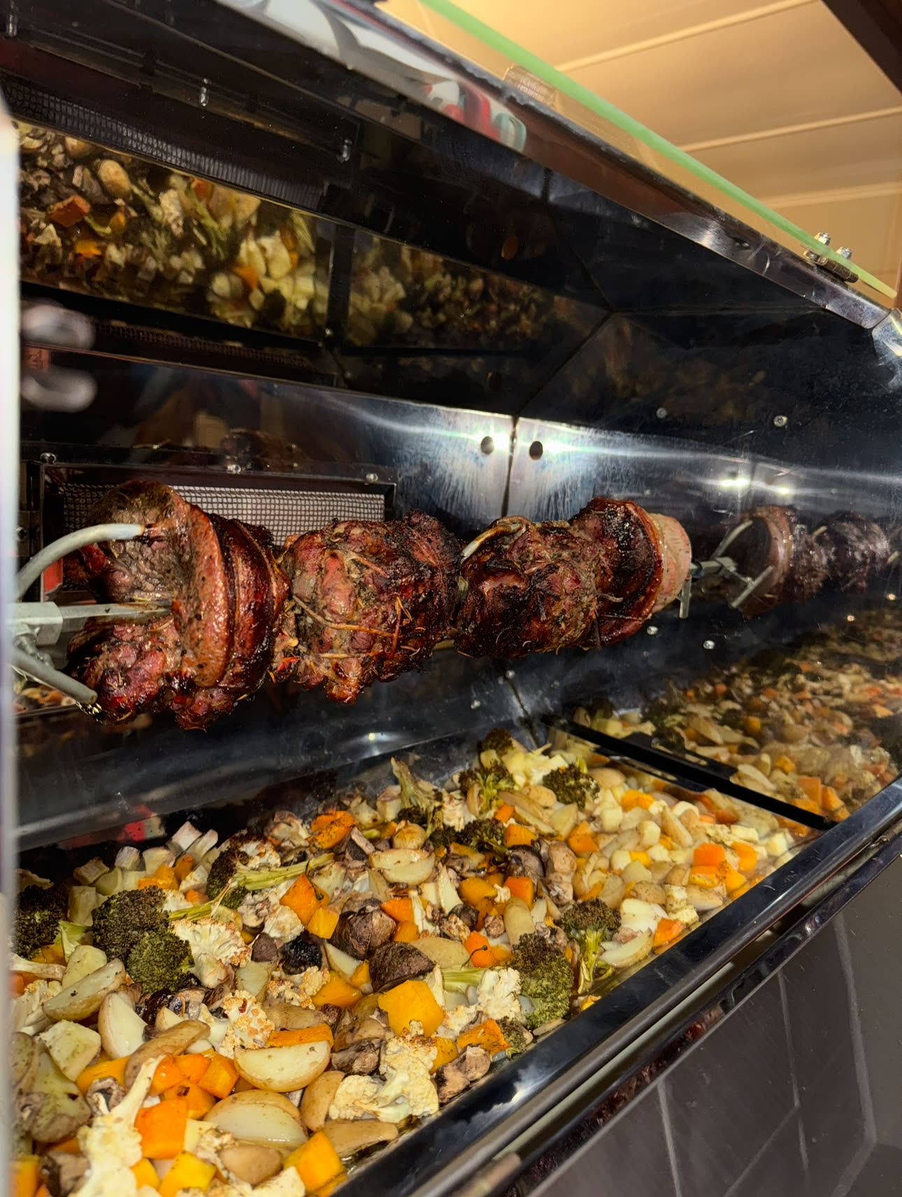 A loaded spit rod of rolled meat above roast vegetables